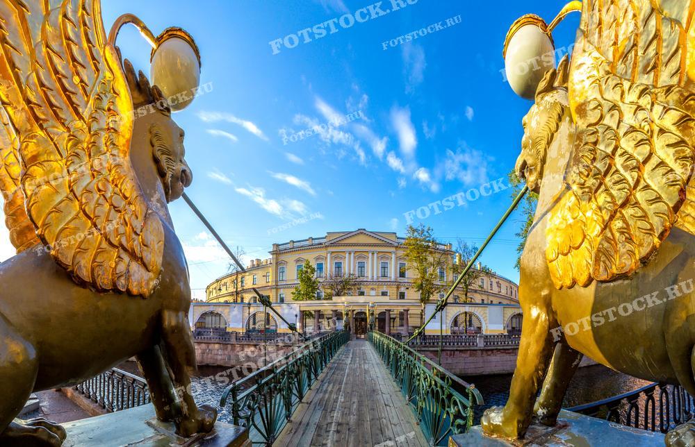 Фотография сделана на Банковском мосту в Санкт-Петербурге и показывает его знаменитых золотых грифонов. Снимок выполнен с такого ракурса, что крылатые существа кажутся огромными, а мост уходит в глубину кадра, создавая перспективу. За спинами грифонов виднеется спокойная городская архитектура под ясным голубым небом с белыми облаками. Это место часто посещается туристами, и фотография могла бы украсить туристические буклеты, посты в социальных сетях о путешествиях или стать уникальным декоративным элементом.